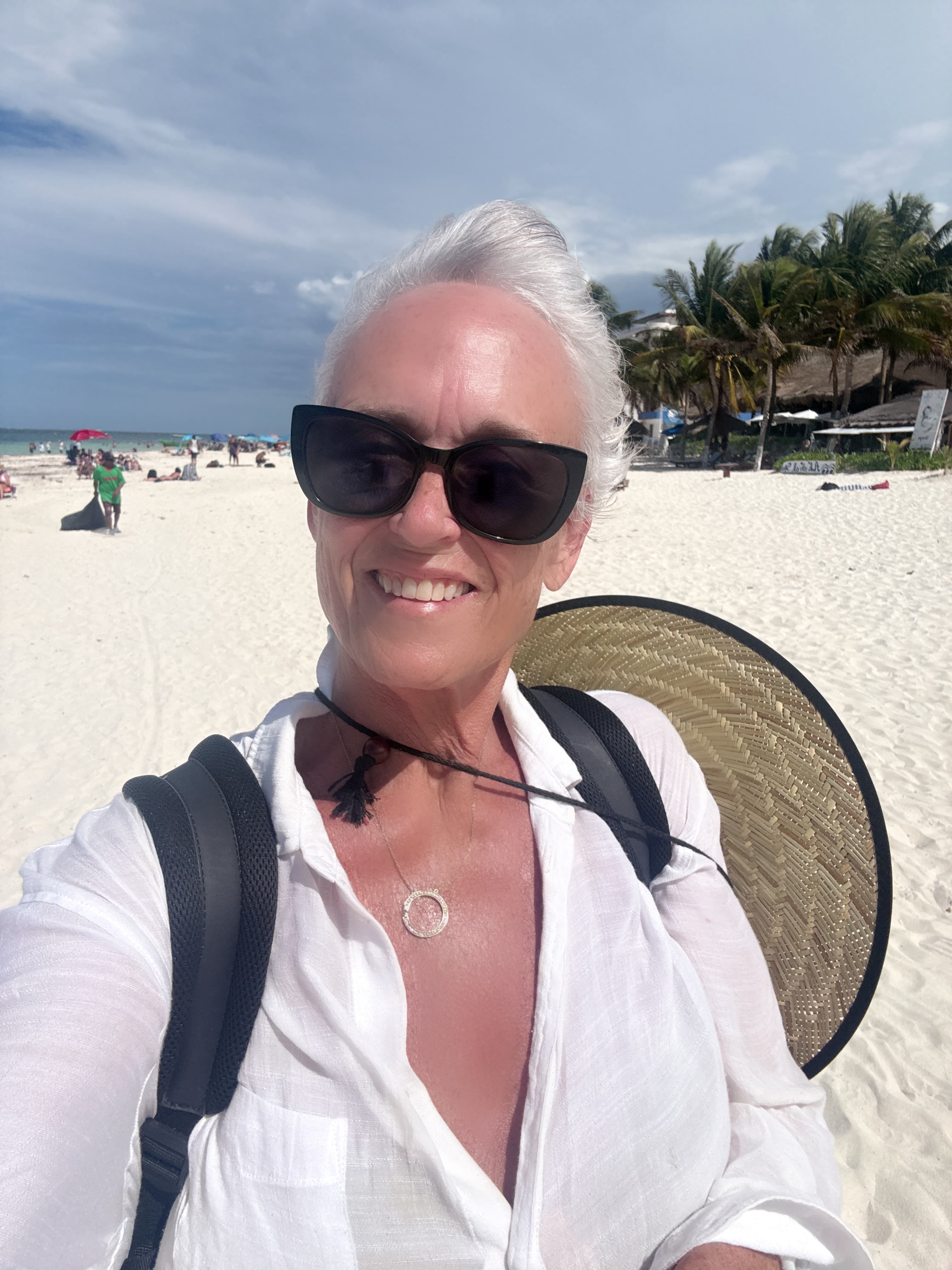 Smiling woman with short white hair and sunglasses on a sunny beach with palm trees.