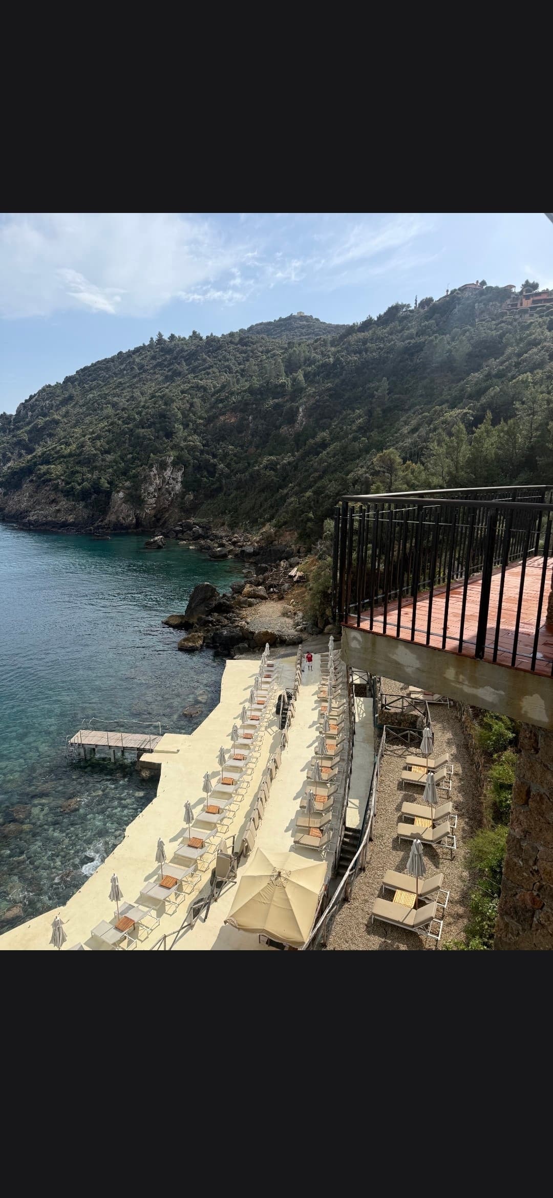 Sun loungers and umbrellas at a beach club overlooking clear water and lush green hills.