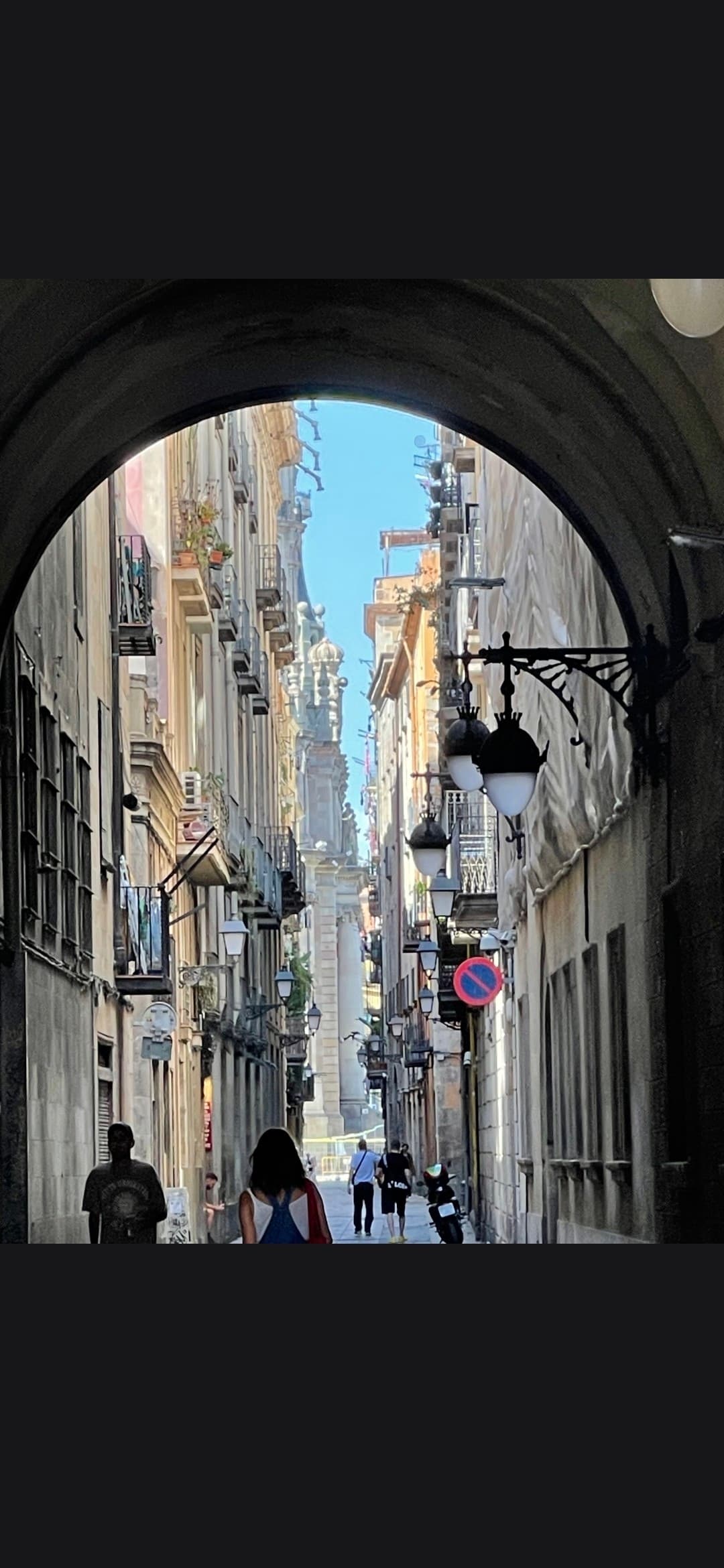 Sunlit narrow street with tall buildings and pedestrians, framed by a dark stone archway.